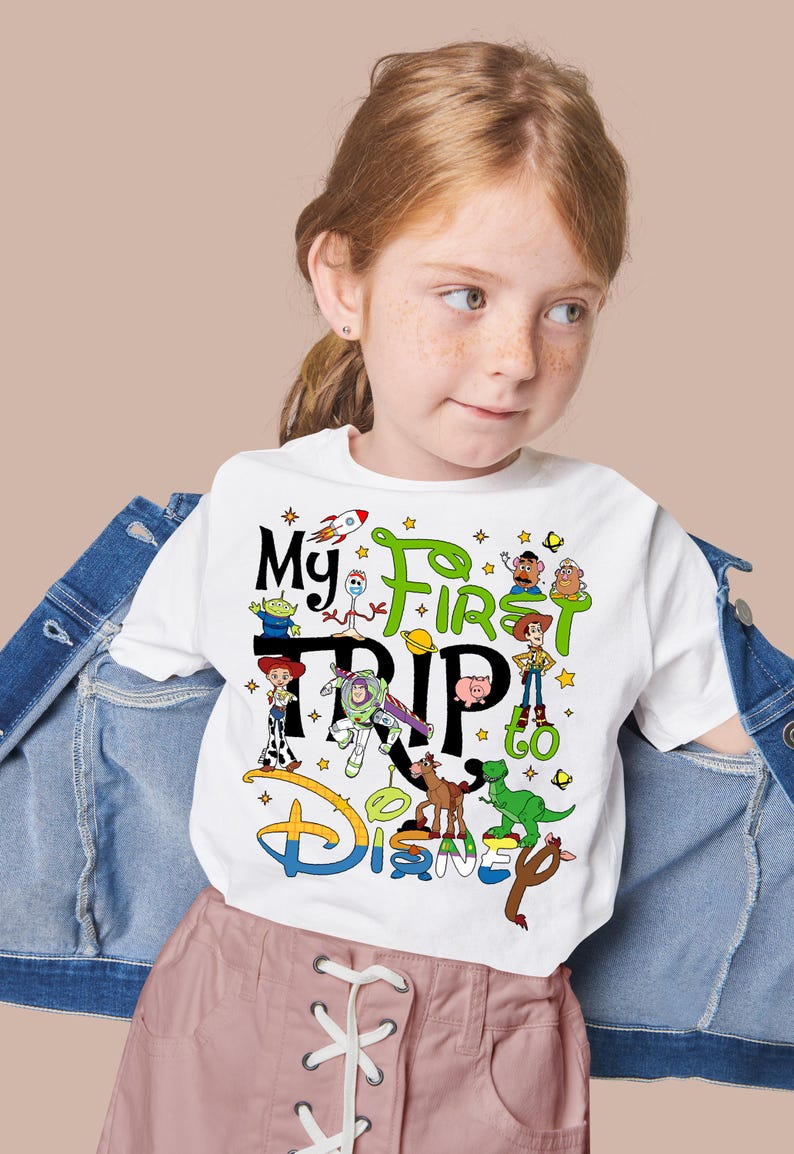 Puede incluir: Camiseta blanca con un gr&aacute;fico colorido con personajes de la pel&iacute;cula Toy Story y el texto "My First Trip to Disney". El dise&ntilde;o incluye un cohete, estrellas y el logotipo de Disney.