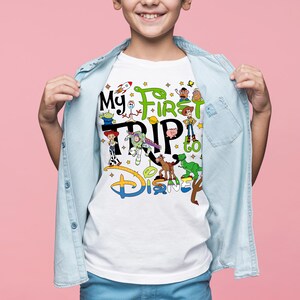 Puede incluir: Camiseta blanca con el texto "My First Trip to Disney" y personajes de dibujos animados coloridos. El dise&ntilde;o incluye personajes de la franquicia Toy Story. La camiseta la lleva un ni&ntilde;o, con una camisa vaquera azul claro abierta.