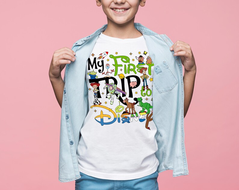 Puede incluir: Camiseta blanca con el texto "My First Trip to Disney" y personajes de dibujos animados coloridos. El dise&ntilde;o incluye personajes de la franquicia Toy Story. La camiseta la lleva un ni&ntilde;o, con una camisa vaquera azul claro abierta.