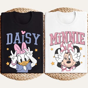 K&ouml;nnte beinhalten: Zwei T-Shirts mit Disney-Figuren. Ein schwarzes T-Shirt mit "DAISY" in blauen Buchstaben und Daisy Duck, die ein Friedenszeichen macht. Das andere ist wei&szlig; mit "MINNIE" in rosa Buchstaben und Minnie Maus, die ein Friedenszeichen macht.
