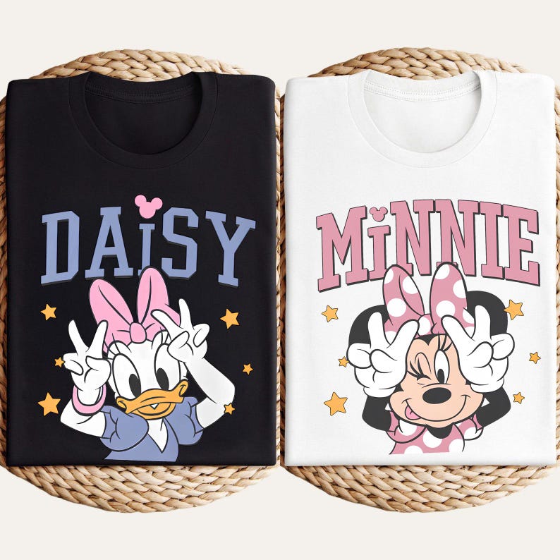 K&ouml;nnte beinhalten: Zwei T-Shirts mit Disney-Figuren. Ein schwarzes T-Shirt mit "DAISY" in blauen Buchstaben und Daisy Duck, die ein Friedenszeichen macht. Das andere ist wei&szlig; mit "MINNIE" in rosa Buchstaben und Minnie Maus, die ein Friedenszeichen macht.