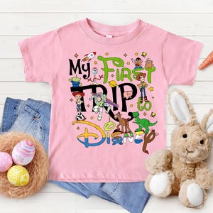 Puede incluir: Una camiseta rosa con el texto "My First Trip to Disney" y varios personajes de dibujos animados. La imagen incluye vaqueros azules, un nido con huevos de Pascua, un conejito de peluche y zanahorias.