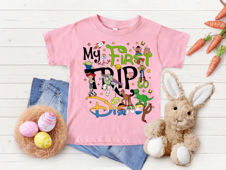 Puede incluir: Una camiseta rosa con el texto "My First Trip to Disney" y varios personajes de dibujos animados. La imagen incluye vaqueros azules, un nido con huevos de Pascua, un conejito de peluche y zanahorias.