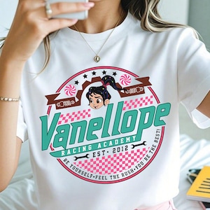 May include: White t-shirt with a circular graphic that reads "Vanellope Racing Academy EST. 2012". The design incorporates a cartoon character, stars, and a pink and white chequered pattern. The shirt also has the text "BE YOURSELF. FEEL THE RUSH. YOU'RE THE BEST!"