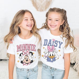 K&ouml;nnte beinhalten: Zwei junge M&auml;dchen tragen wei&szlig;e T-Shirts mit Zeichentrickfiguren. Eines zeigt "MINNIE" in Rosa, das andere "DAISY" in Blau. Beide Shirts zeigen Zeichentrickfiguren und Friedenszeichen, ideal f&uuml;r Disney-Fans.