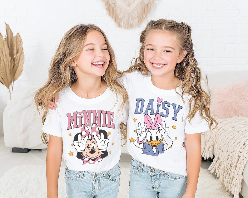 K&ouml;nnte beinhalten: Zwei junge M&auml;dchen tragen wei&szlig;e T-Shirts mit Zeichentrickfiguren. Eines zeigt "MINNIE" in Rosa, das andere "DAISY" in Blau. Beide Shirts zeigen Zeichentrickfiguren und Friedenszeichen, ideal f&uuml;r Disney-Fans.
