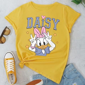 K&ouml;nnte beinhalten: Senfgelbes T-Shirt mit Daisy Duck-Grafik und dem Wort "DAISY" in Blau. Das Bild zeigt blaue Jeansshorts, gelbe Turnschuhe und eine Sonnenbrille. Ein Strau&szlig; gelber Blumen befindet sich im Hintergrund.