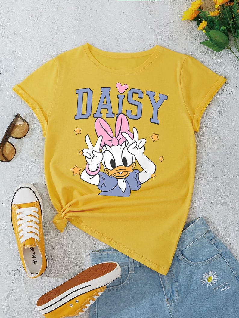 K&ouml;nnte beinhalten: Senfgelbes T-Shirt mit Daisy Duck-Grafik und dem Wort "DAISY" in Blau. Das Bild zeigt blaue Jeansshorts, gelbe Turnschuhe und eine Sonnenbrille. Ein Strau&szlig; gelber Blumen befindet sich im Hintergrund.