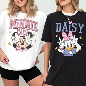 K&ouml;nnte beinhalten: Zwei T-Shirts mit Disney-Figuren. Eines ist wei&szlig; mit "Minnie" in Rosa und einer Minnie Mouse-Grafik. Das andere ist schwarz mit "Daisy" in Blau und einer Daisy Duck-Grafik. Beide Shirts haben Peace-Zeichen.