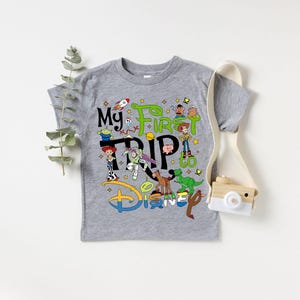 Puede incluir: Una camiseta gris de manga corta con el texto "My First Trip to Disney" y personajes de dibujos animados coloridos. El dise&ntilde;o incluye un cohete, estrellas y el logotipo de Disney en azul, verde y amarillo.