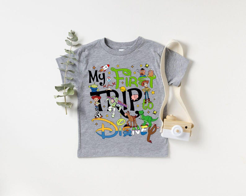 Puede incluir: Una camiseta gris de manga corta con el texto "My First Trip to Disney" y personajes de dibujos animados coloridos. El dise&ntilde;o incluye un cohete, estrellas y el logotipo de Disney en azul, verde y amarillo.