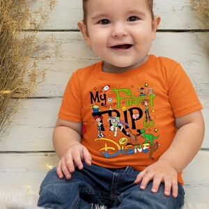 Puede incluir: Una camiseta naranja con el texto "My First Trip to Disney" y varios personajes de Toy Story. El beb&eacute; lleva vaqueros azules. La imagen muestra a un beb&eacute; sonriendo.