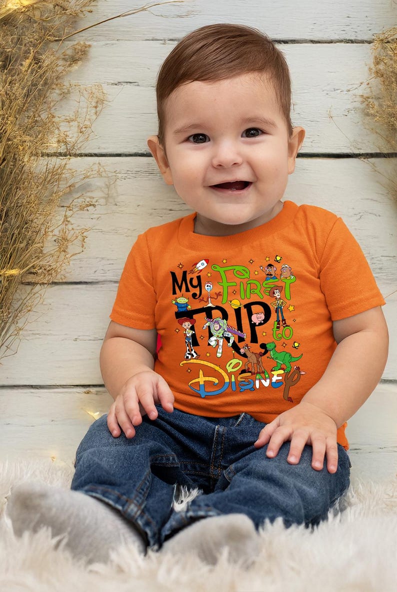 Puede incluir: Una camiseta naranja con el texto "My First Trip to Disney" y varios personajes de Toy Story. El beb&eacute; lleva vaqueros azules. La imagen muestra a un beb&eacute; sonriendo.