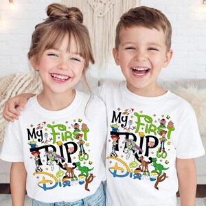 Puede incluir: Dos ni&ntilde;os con camisetas blancas con un gr&aacute;fico colorido. Las camisetas dicen "My First Trip to Disney" con personajes de dibujos animados. El dise&ntilde;o incluye un cohete, estrellas y el logo de Disney.