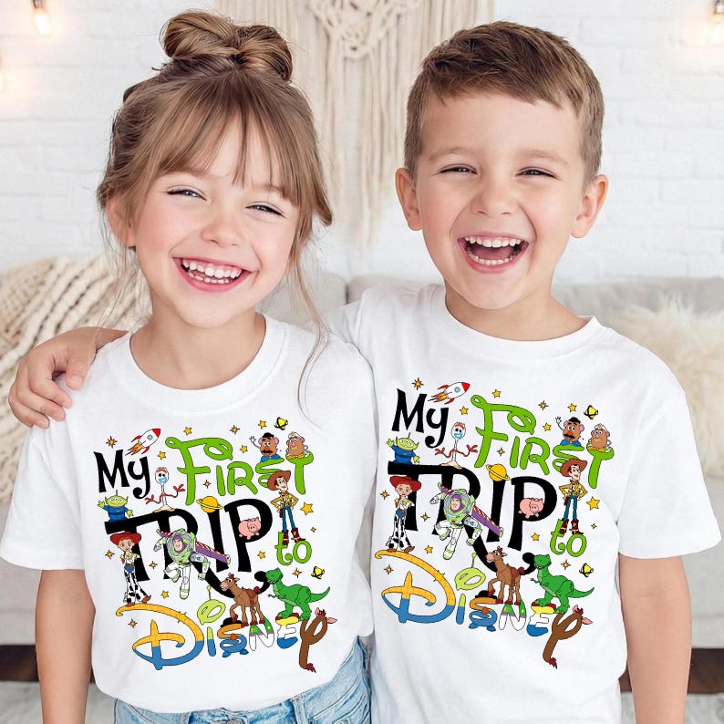 Puede incluir: Dos ni&ntilde;os con camisetas blancas con un gr&aacute;fico colorido. Las camisetas dicen "My First Trip to Disney" con personajes de dibujos animados. El dise&ntilde;o incluye un cohete, estrellas y el logo de Disney.