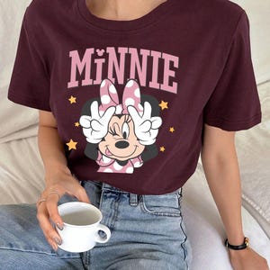 K&ouml;nnte beinhalten: Burgunderfarbenes T-Shirt mit einem Minnie Mouse-Motiv. Die Grafik zeigt das Wort "MINNIE" in Rosa &uuml;ber einem Cartoon-Bild von Minnie Mouse mit einer gepunkteten Schleife und H&auml;nden, die Friedenszeichen machen. Goldene Sterne sind um das Bild verstreut.