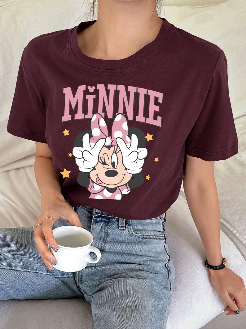 K&ouml;nnte beinhalten: Burgunderfarbenes T-Shirt mit einem Minnie Mouse-Motiv. Die Grafik zeigt das Wort "MINNIE" in Rosa &uuml;ber einem Cartoon-Bild von Minnie Mouse mit einer gepunkteten Schleife und H&auml;nden, die Friedenszeichen machen. Goldene Sterne sind um das Bild verstreut.