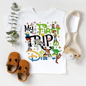 Puede incluir: Camiseta blanca con un gr&aacute;fico colorido de los personajes de Toy Story y el texto "My First Trip to Disney". El dise&ntilde;o incluye un cohete, estrellas y el logotipo de Disney. La camiseta se combina con zapatos de cuero marrones.