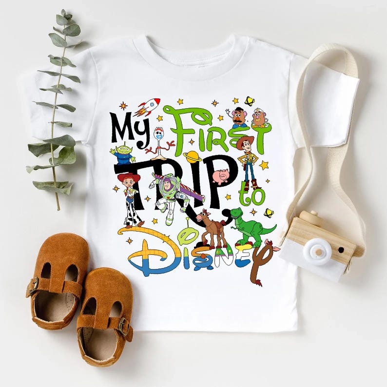 Puede incluir: Camiseta blanca con un gr&aacute;fico colorido de los personajes de Toy Story y el texto "My First Trip to Disney". El dise&ntilde;o incluye un cohete, estrellas y el logotipo de Disney. La camiseta se combina con zapatos de cuero marrones.