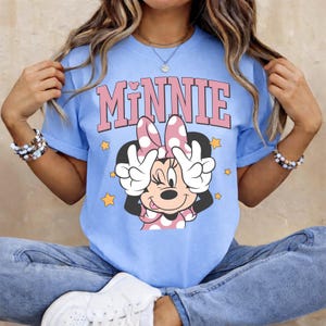 K&ouml;nnte beinhalten: Hellblaues T-Shirt mit einem Minnie Mouse-Motiv. Das Wort "MINNIE" steht in rosa Blockbuchstaben &uuml;ber einer Cartoon-Minnie-Maus, die das Friedenszeichen zeigt. Gelbe Sterne umgeben die Figur. Das T-Shirt hat kurze &Auml;rmel.