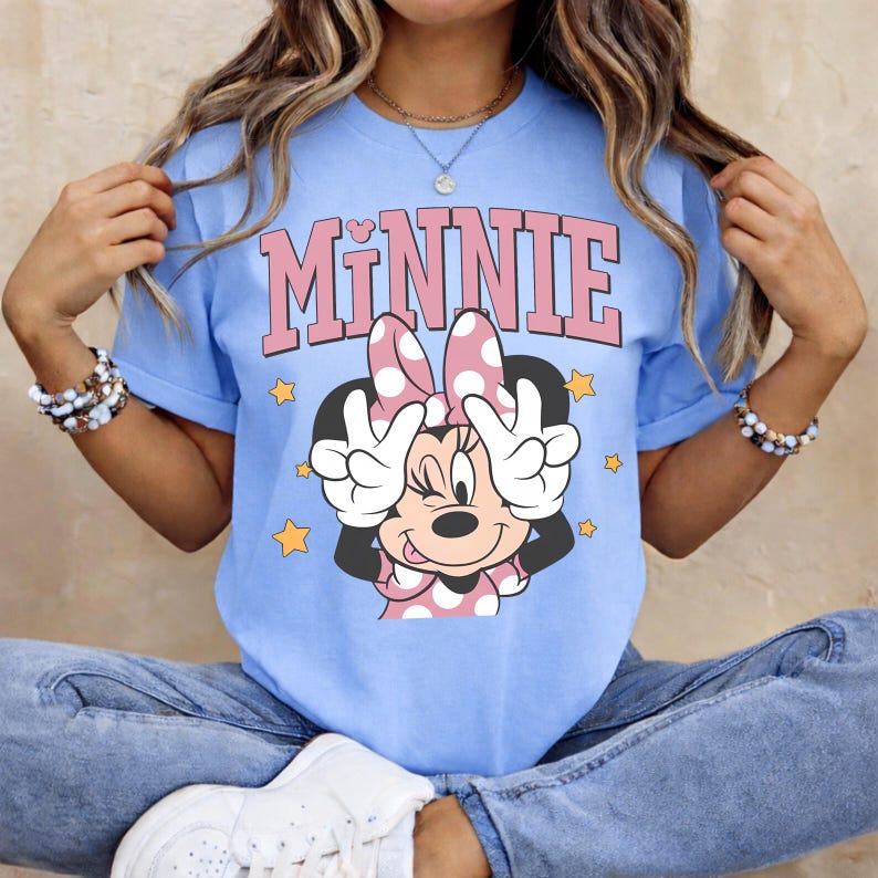 K&ouml;nnte beinhalten: Hellblaues T-Shirt mit einem Minnie Mouse-Motiv. Das Wort "MINNIE" steht in rosa Blockbuchstaben &uuml;ber einer Cartoon-Minnie-Maus, die das Friedenszeichen zeigt. Gelbe Sterne umgeben die Figur. Das T-Shirt hat kurze &Auml;rmel.