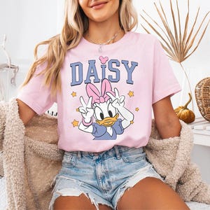 K&ouml;nnte beinhalten: Rosafarbenes T-Shirt mit dem Wort "DAISY" in Blau, einem rosa Herz und einem Cartoon-Bild von Daisy Duck, die Friedenszeichen zeigt. Das T-Shirt wird mit Jeansshorts getragen.