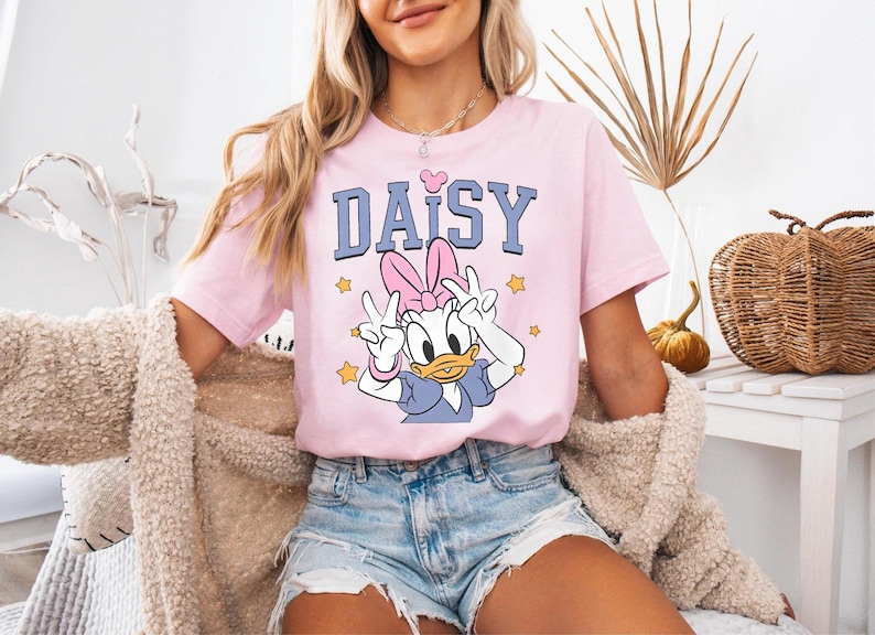K&ouml;nnte beinhalten: Rosafarbenes T-Shirt mit dem Wort "DAISY" in Blau, einem rosa Herz und einem Cartoon-Bild von Daisy Duck, die Friedenszeichen zeigt. Das T-Shirt wird mit Jeansshorts getragen.