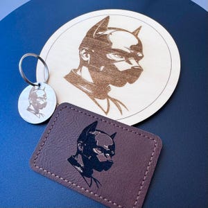 May include: A circular light wood coaster, a silver metal circular keychain, and a rectangular brown faux leather patch. Each item features a laser-engraved or printed graphic of a person wearing a pointed-ear hood or mask in profile view.