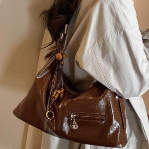 Pode incluir: Uma bolsa hobo castanho-escura com uma textura brilhante e enrugada. A bolsa apresenta um bolso frontal horizontal com fecho de correr e puxador prateado, e um pendente decorativo composto por contas de madeira castanha e um anel de metal prateado.