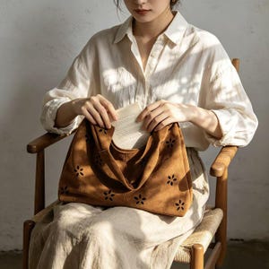 May include: A brown suede tote bag with a floral cut-out design. The bag has a soft, slouchy shape and a single handle. The bag is held open, revealing a book inside. The bag is a stylish accessory.