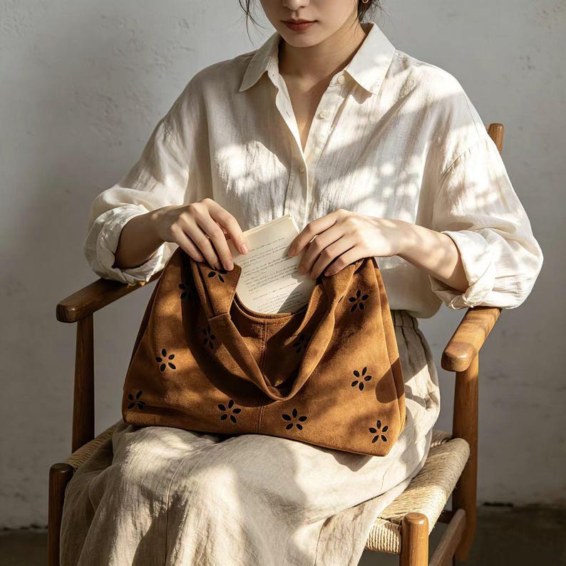 May include: A brown suede tote bag with a floral cut-out design. The bag has a soft, slouchy shape and a single handle. The bag is held open, revealing a book inside. The bag is a stylish accessory.