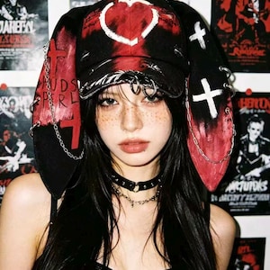 May include: Black and red hat with long ear flaps, a heart design, and white crosses. The hat is adorned with metal rings and chains. The person is wearing a black choker necklace.