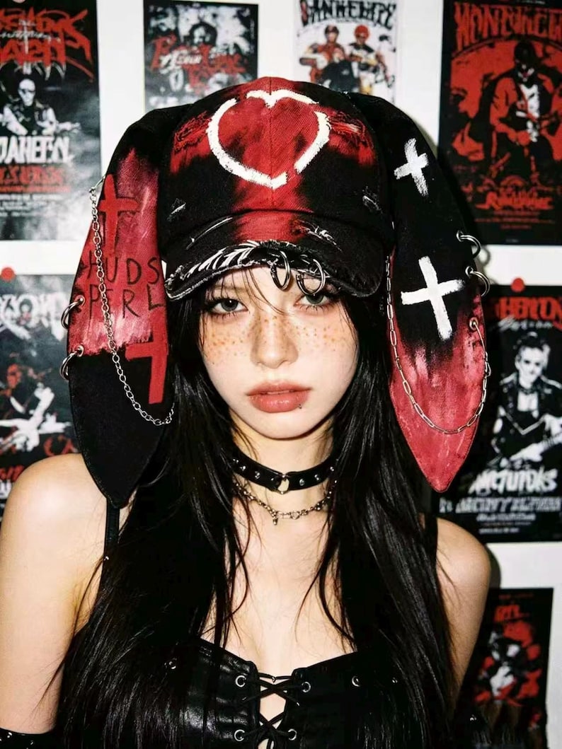 May include: Black and red hat with long ear flaps, a heart design, and white crosses. The hat is adorned with metal rings and chains. The person is wearing a black choker necklace.