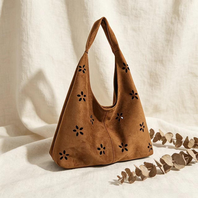 May include: A brown suede hobo bag with a single shoulder strap. The bag features a series of small, flower-shaped cutouts across the front. The bag is set against a neutral background, with a sprig of dried leaves in the lower right corner.