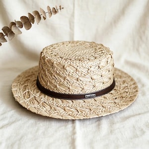 Puede incluir: Un sombrero de paja de color tostado claro con copa plana y ala ancha, que presenta una banda de cuero marrón oscuro alrededor de la base de la copa. Una pequeña placa metálica rectangular de tono plateado está fijada a la banda con el texto "LAVISH".
