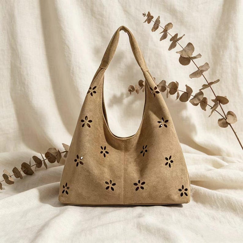 May include: A tan suede shoulder bag with a floral cut-out design. The bag has a single handle and a curved shape. The bag is sitting on a white surface with dried foliage in the background.
