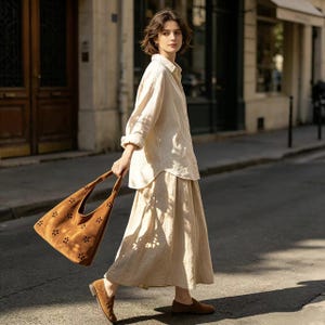 May include: A woman wearing a cream-colored linen shirt and skirt, paired with brown suede loafers. She carries a brown suede tote bag with floral cutouts. The outfit is suitable for a casual, everyday look.