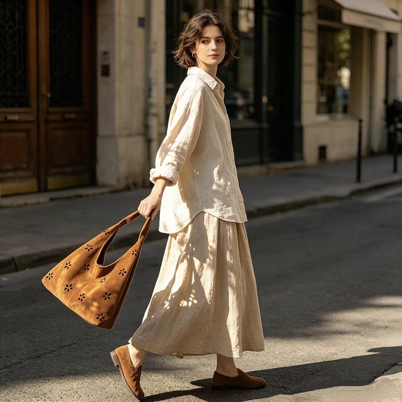 May include: A woman wearing a cream-colored linen shirt and skirt, paired with brown suede loafers. She carries a brown suede tote bag with floral cutouts. The outfit is suitable for a casual, everyday look.