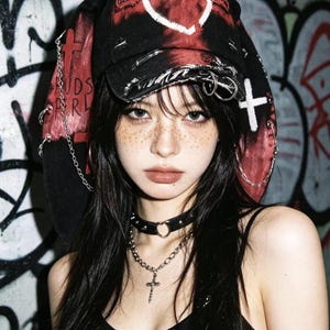 May include: A black and red hat with a heart design and a cross. The hat has chains and metal rings. The person is wearing a black choker with a silver cross necklace. The background is a graffiti wall.