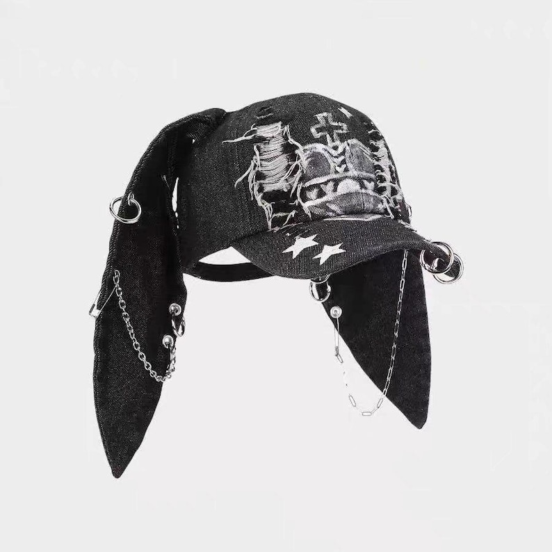 May include: Dark gray denim cap with long, pointed ear flaps. The cap features a distressed design with a white embroidered crown and star accents. Silver rings and chains dangle from the ear flaps and brim, adding a punk rock aesthetic.