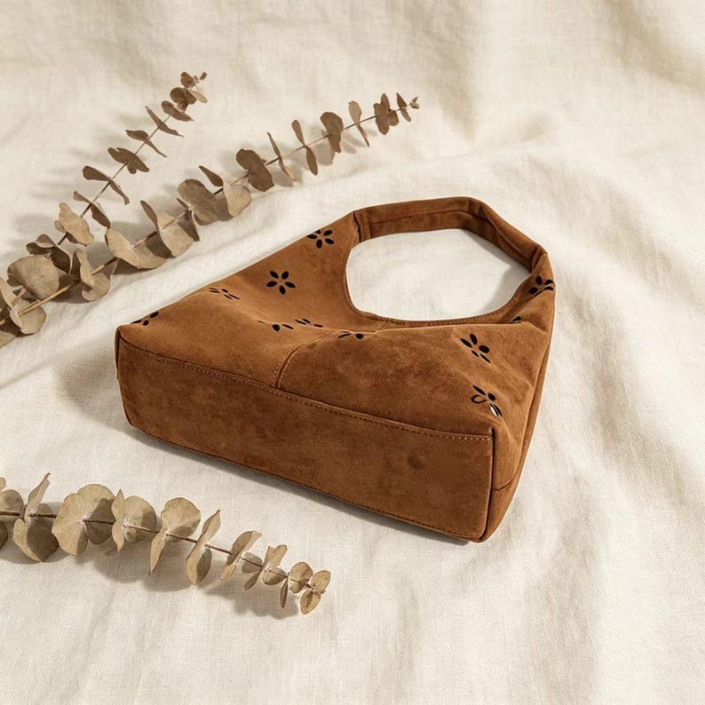 May include: A brown suede shoulder bag with a rectangular base and a curved top handle. The bag features a floral cut-out pattern and is set against a neutral background with dried foliage.