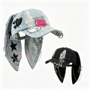 Puede incluir: Una gorra de béisbol de mezclilla azul claro desgastada con un gráfico de corazón rosa con alas, el texto Hudson y solapas laterales largas con estrellas negras y cadenas plateadas. Una segunda gorra negra desgastada con solapas y cadenas.