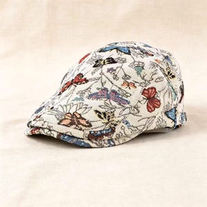 May include: A flat cap featuring a cream-colored base with a woven pattern of multi-colored butterflies and floral elements. The cap has a structured brim and a rounded crown, showcasing a detailed tapestry-style fabric with red, blue, and black accents.
