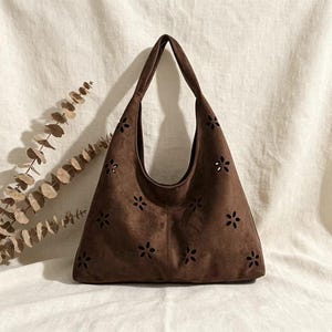 May include: A brown suede shoulder bag with a floral cut-out design. The bag has a large, open top and a single shoulder strap. The bag is made of a soft, suede-like material and is a medium size.