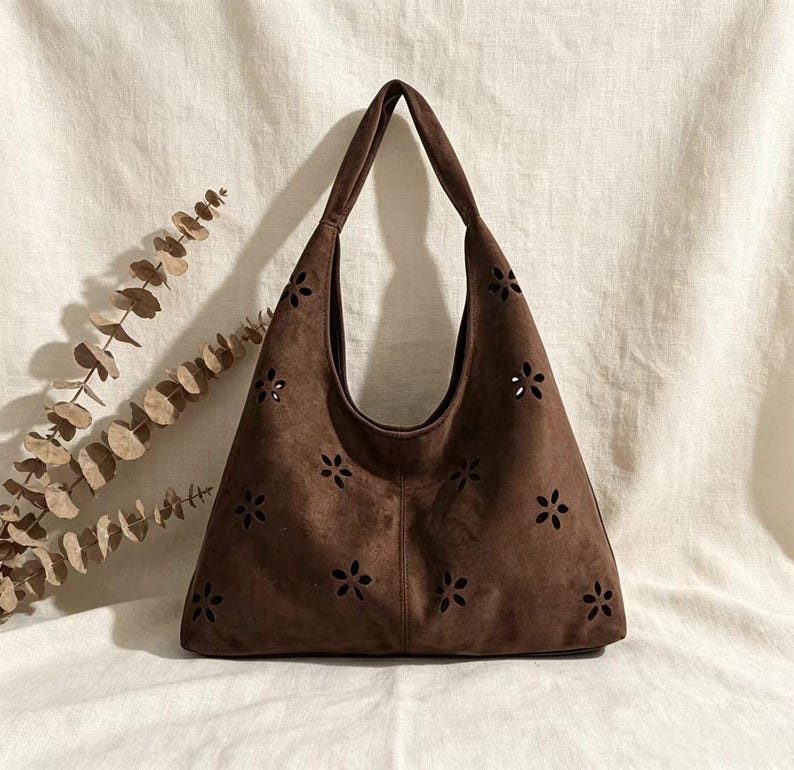 May include: A brown suede shoulder bag with a floral cut-out design. The bag has a large, open top and a single shoulder strap. The bag is made of a soft, suede-like material and is a medium size.