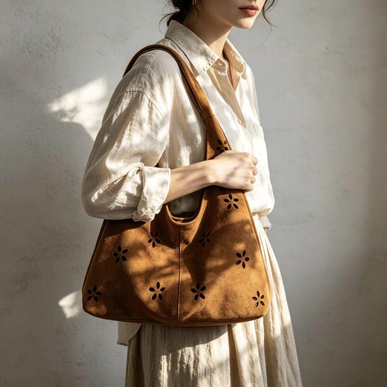 May include: A brown suede tote bag with a floral cut-out design. The bag has a wide shoulder strap and a spacious interior. The bag is held by a person wearing a cream-colored shirt and skirt. The bag is approximately 15 inches wide and 12 inches tall.