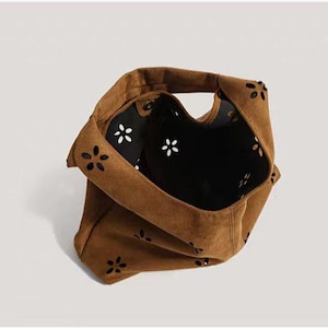 May include: A brown suede tote bag with a floral cut-out design. The bag has a single handle and a black interior with white flower accents. The bag is made of a soft, textured material.