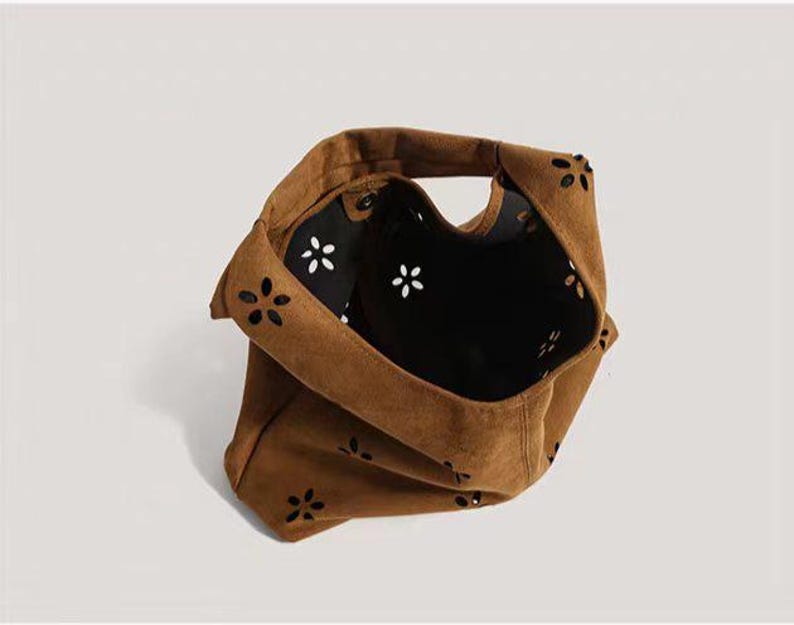May include: A brown suede tote bag with a floral cut-out design. The bag has a single handle and a black interior with white flower accents. The bag is made of a soft, textured material.