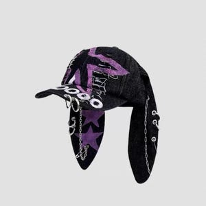 May include: Black denim cap with purple star and zigzag designs. The cap features silver hoop and chain accents, and long, ear-like flaps. The word "2000" is embroidered on the brim.