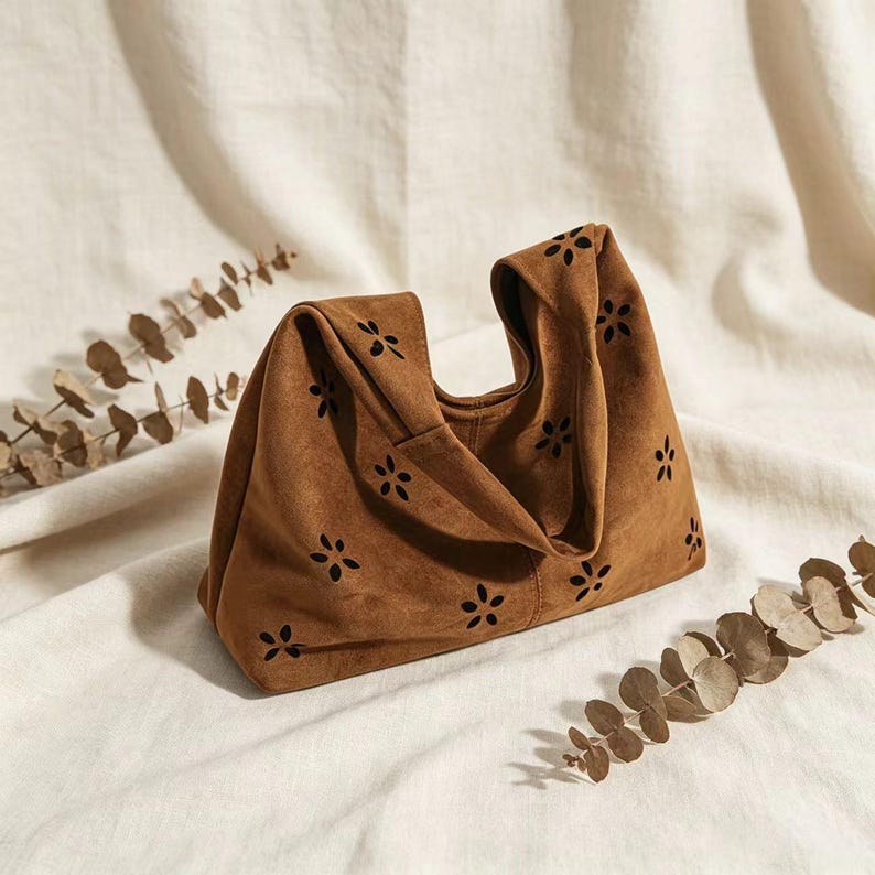 May include: A brown suede handbag with a unique design. The bag features a folded top and is adorned with small, flower-shaped cutouts. The bag is set against a neutral background with dried foliage, highlighting its texture and style.